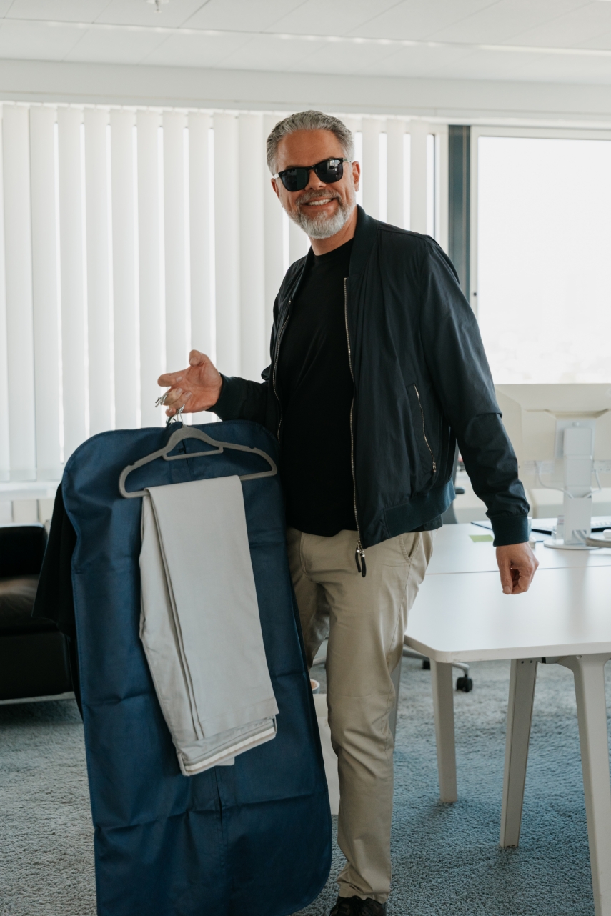 Hans Mantell steht lächelnd mit Sonnenbrille im Büro und hält einen blauen Kleidersack mit einem Anzug auf einem Kleiderbügel.